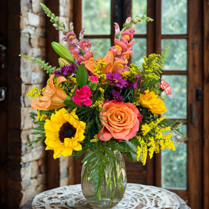Mixed bouquet in a clear glass vase with sunflowers, roses, lilies, and snapdragons