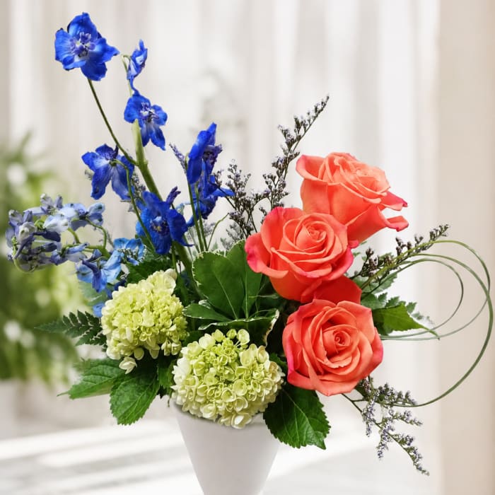 Bouquet of coral roses, blue flowers, and pale green hydrangeas in a white vase