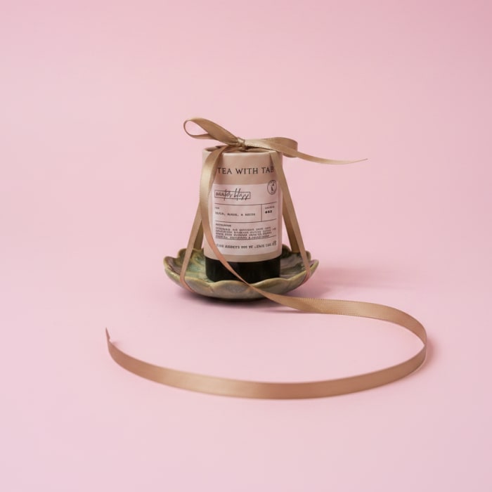 Tea bottle tied with a gold ribbon on a small dish against a pink background