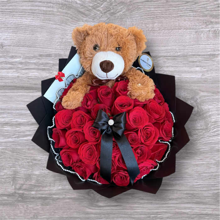 Teddy bear sitting on a bouquet of red roses with a black ribbon