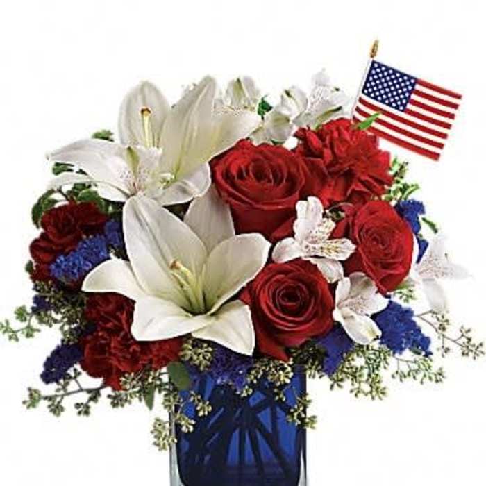 Patriotic arrangement of red roses and white lilies in a blue glass vase with a small American flag.
