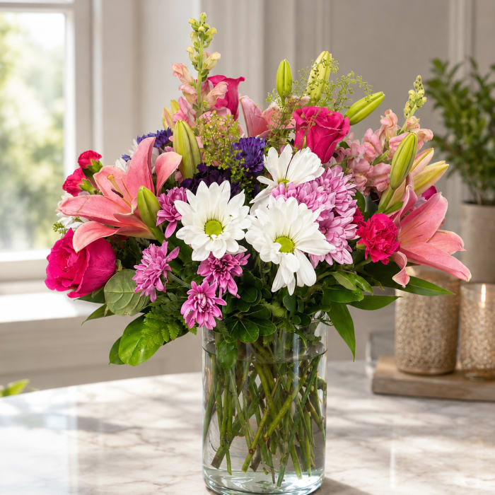Mixed bouquet of pink lilies, roses, daisies, and purple blooms in a glass vase