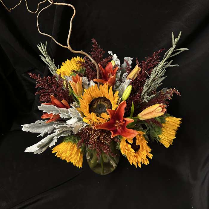 Sunflowers and orange lilies arranged in a glass vase with curly branches