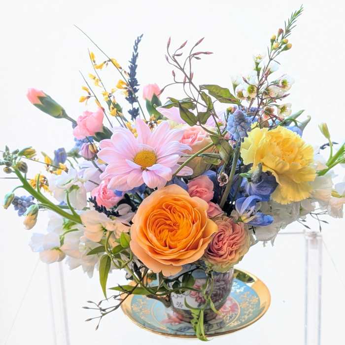 Pastel arrangement of roses, daisies, and mixed blooms in a patterned teacup on a matching saucer