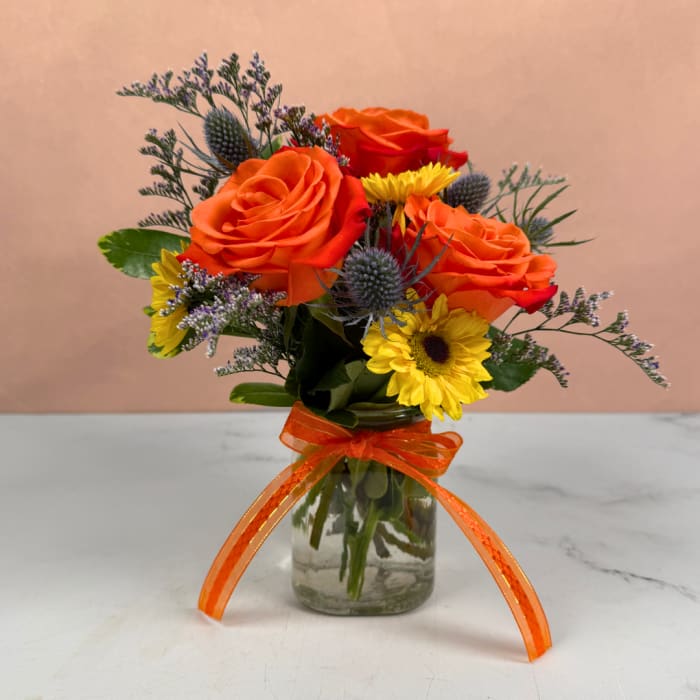 Orange roses and yellow flowers in a glass vase with an orange ribbon