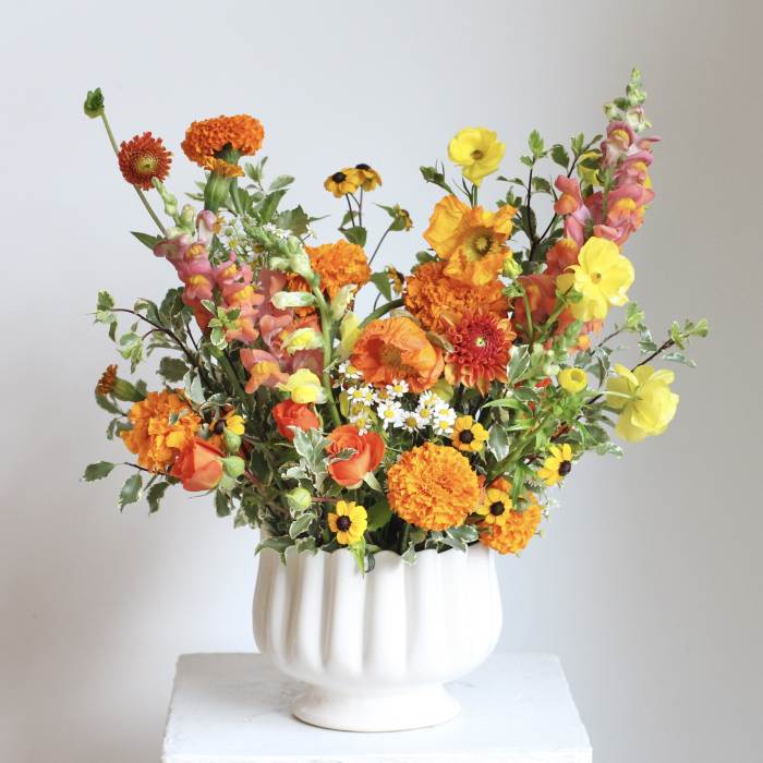 Bright mixed bouquet in a white pedestal vase with orange, yellow, and pink blooms.