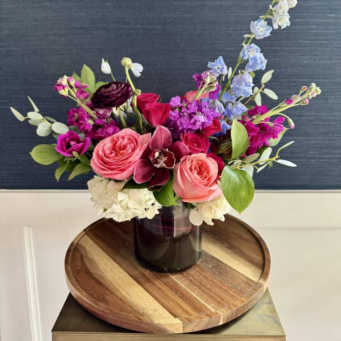 Mixed arrangement of pink roses, white hydrangeas, and purple blooms in a dark glass vase on a wooden tray