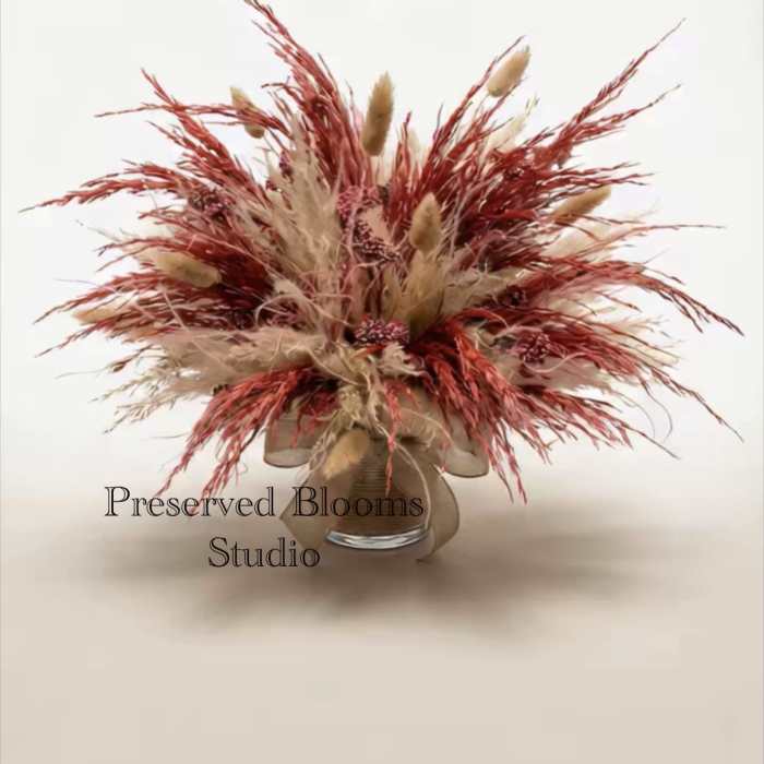 Preserved arrangement of tan and rust dried grasses and seed clusters in a clear glass vase.