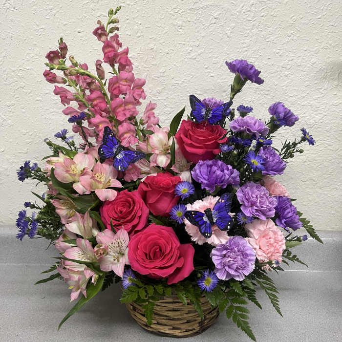 Basket arrangement of pink roses, purple carnations, and pink snapdragons with butterfly picks