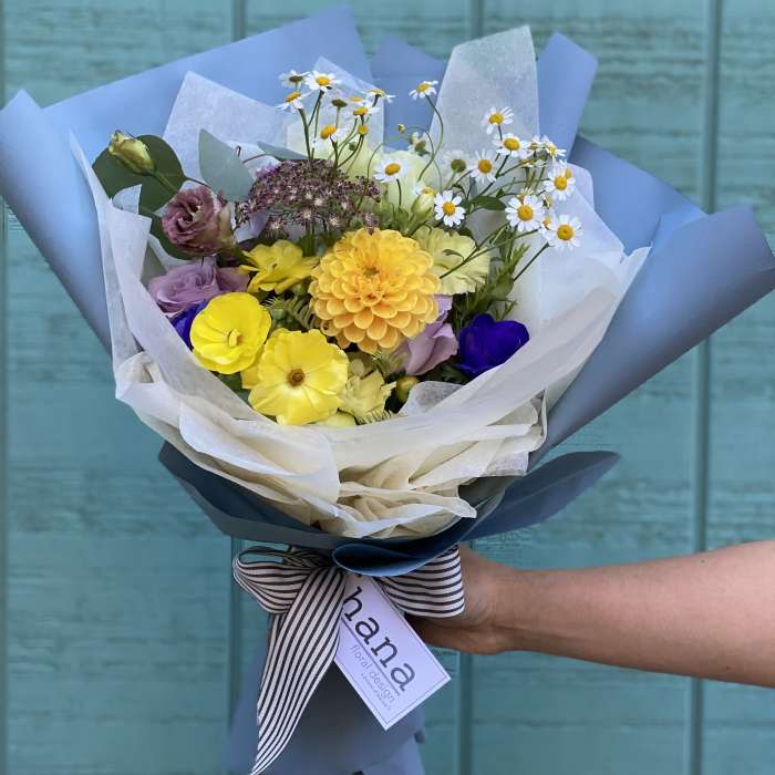 Hand-tied bouquet of yellow, purple, and white flowers wrapped in blue paper