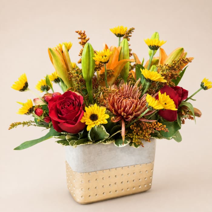 Mixed bouquet with red roses, yellow blooms, and orange lilies in a gold vase