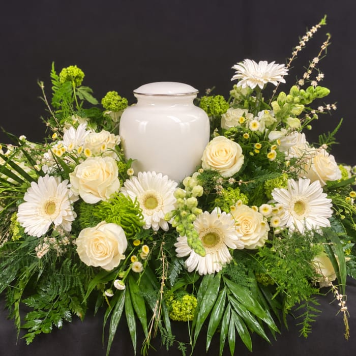 White urn surrounded by cream roses and white daisies in a green floral arrangement