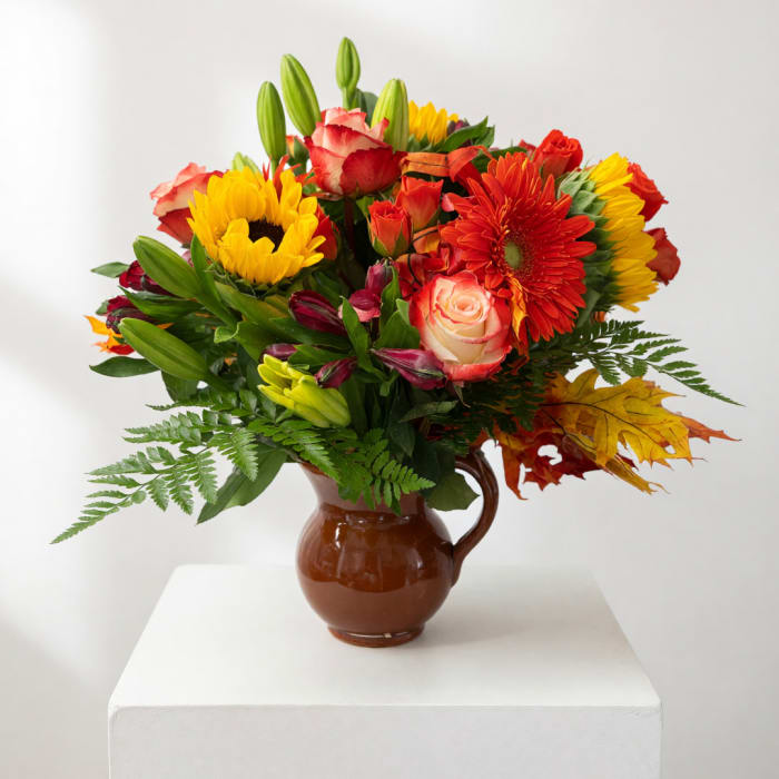 Mixed bouquet of sunflowers, roses, and lilies in a brown pitcher vase