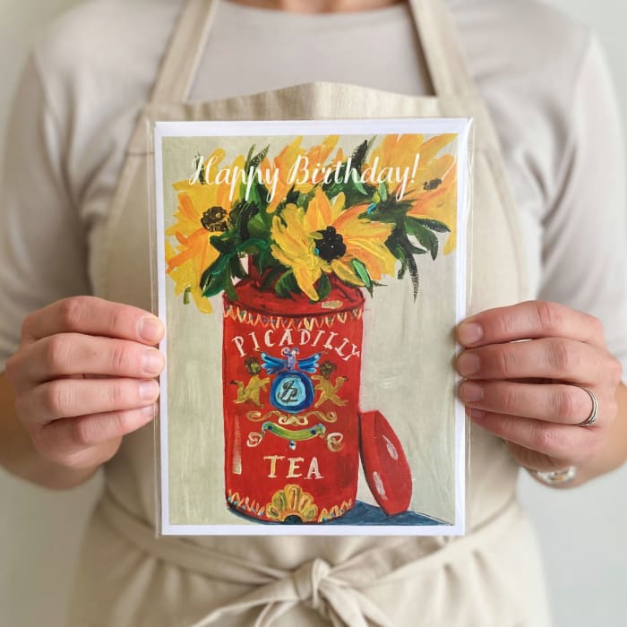 Person holding a birthday card with a painted flower-filled tea tin