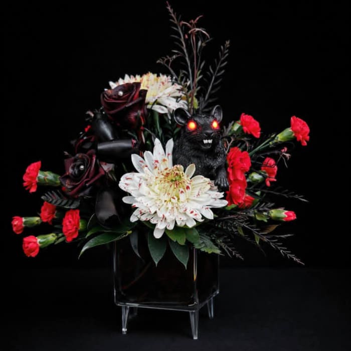 Dark floral arrangement with a glowing rat figurine in a glass vase