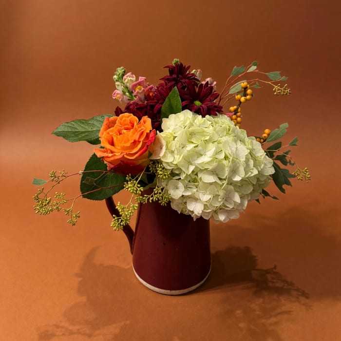 Mixed bouquet with orange rose, white hydrangea, and burgundy blooms in a red pitcher