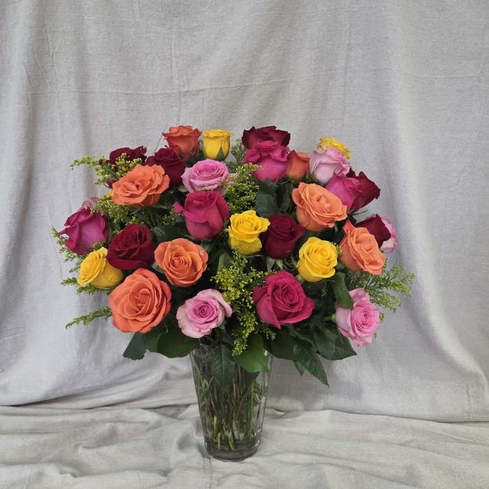 Tall clear vase filled with mixed red, pink, orange, and yellow roses with small yellow filler flowers