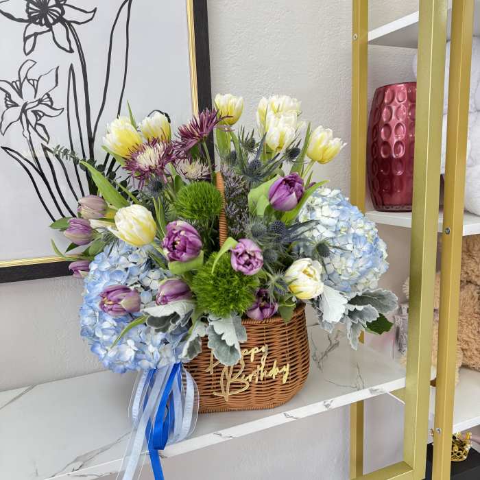Basket arrangement of purple, white, and blue flowers with a birthday ribbon