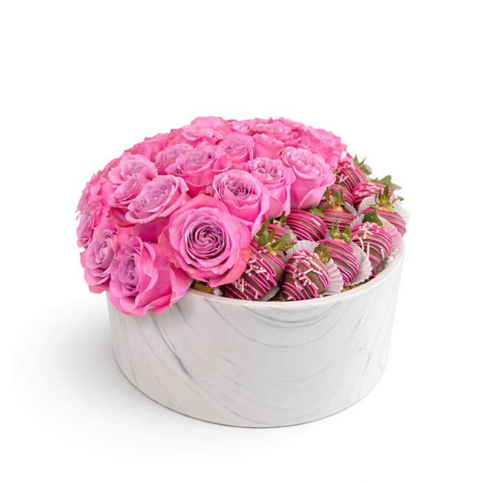 Round white box filled with pink roses and chocolate-covered strawberries drizzled with pink and white.