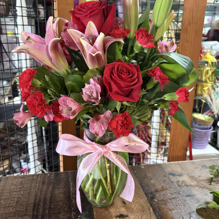 Bouquet of red roses, pink lilies, and carnations in a glass vase with a pink ribbon