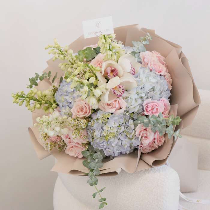 Pastel bouquet of pink roses, blue hydrangeas, and white orchids wrapped in beige paper