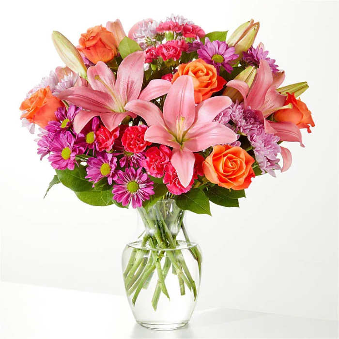 Mixed bouquet of pink lilies, orange roses, and purple daisies in a glass vase