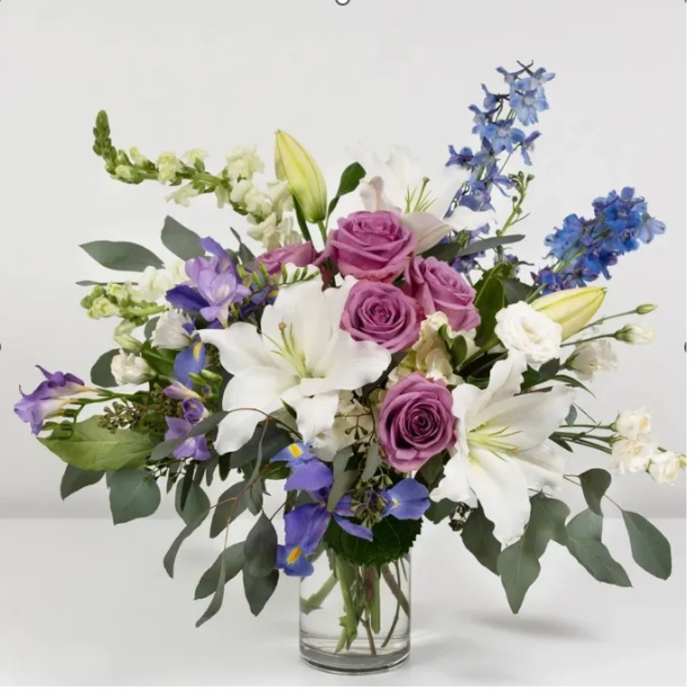 Arrangement of lavender roses, white lilies, and blue flowers in a clear glass vase