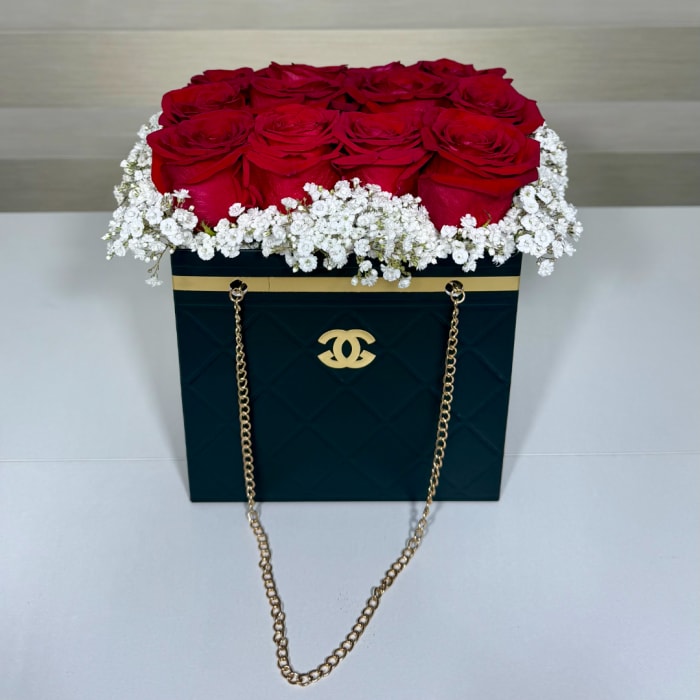 Red roses arranged in a black box with white baby's breath and gold chain handles.