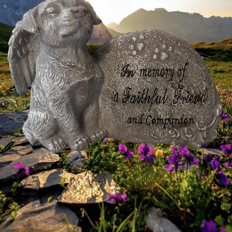 A memorial dog statue beside an engraved stone in a mountain meadow.