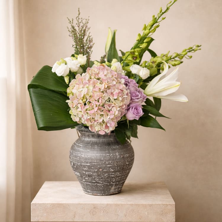 Asymmetrical arrangement of blush hydrangea, lavender roses, white lilies, and broad green leaves in a gray ceramic vase