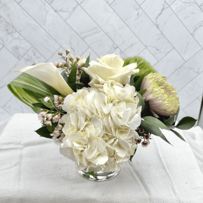 Low white floral arrangement with hydrangea, roses, and calla lilies in a clear glass vase