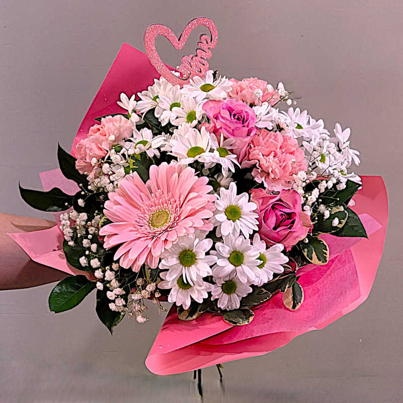 Hand-tied bouquet of pink gerbera, roses, and carnations with white daisies wrapped in pink paper and a heart pick.