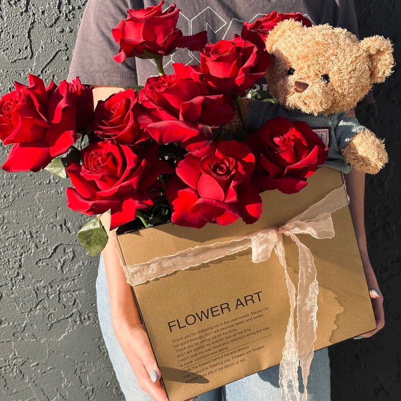 Box of red roses with a small teddy bear held in someone’s arms