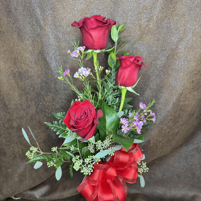 Three red roses with purple filler in a tall vase, tied with a red ribbon