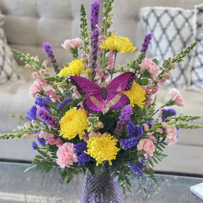 Bouquet of yellow, pink, and purple flowers in a purple vase with a butterfly decoration.