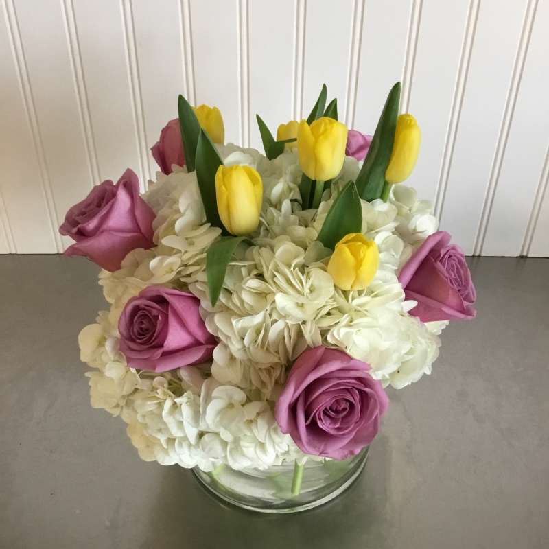 Compact arrangement of yellow tulips, lavender roses, and white hydrangeas in a clear glass vase