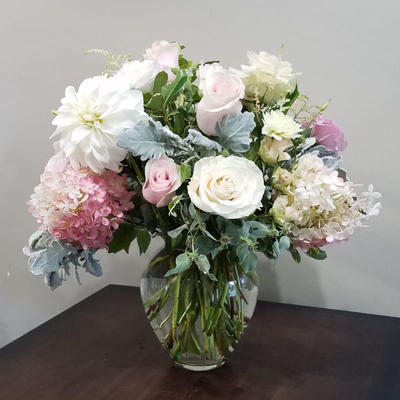 Arrangement of white and blush roses, hydrangeas, and a dahlia in a clear glass vase on a dark table