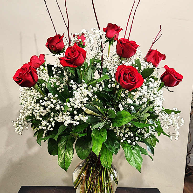 Tall arrangement of red roses and white filler flowers in a clear glass vase on a dark table