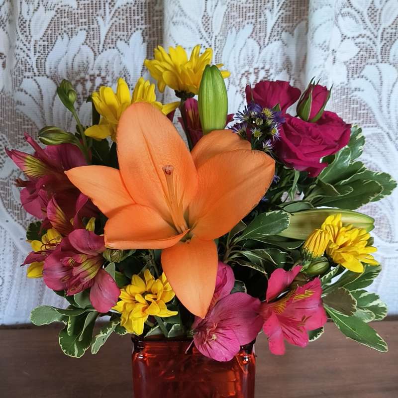 Mixed bouquet with orange lily, pink roses, yellow daisies, and a red vase