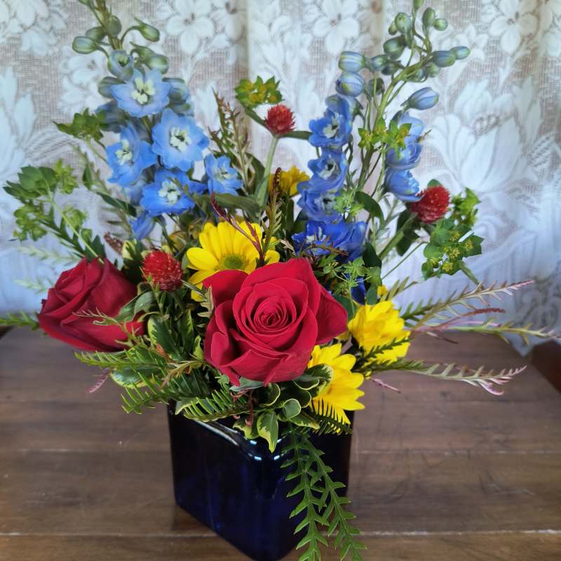 Mixed bouquet of red roses, blue flowers, and yellow daisies in a dark vase