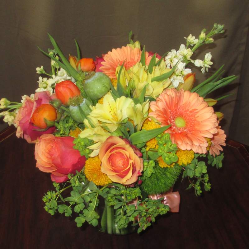 Low glass vase of orange gerberas, roses, and mixed yellow flowers in a compact arrangement