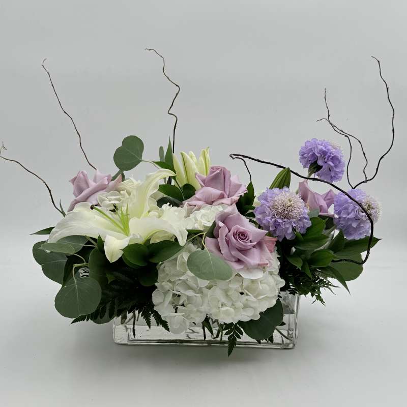 Lavender roses and white lilies arranged with purple blooms in a clear rectangular vase.