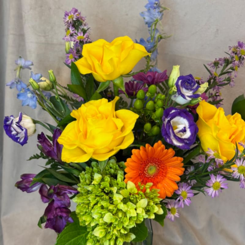 Mixed bouquet with yellow roses, orange gerbera daisy, and purple blooms in a glass vase