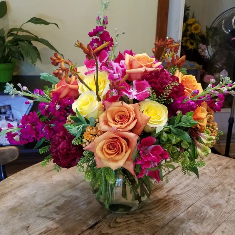 Round glass vase of orange, yellow, and fuchsia flowers including roses and lilies on a wooden table.