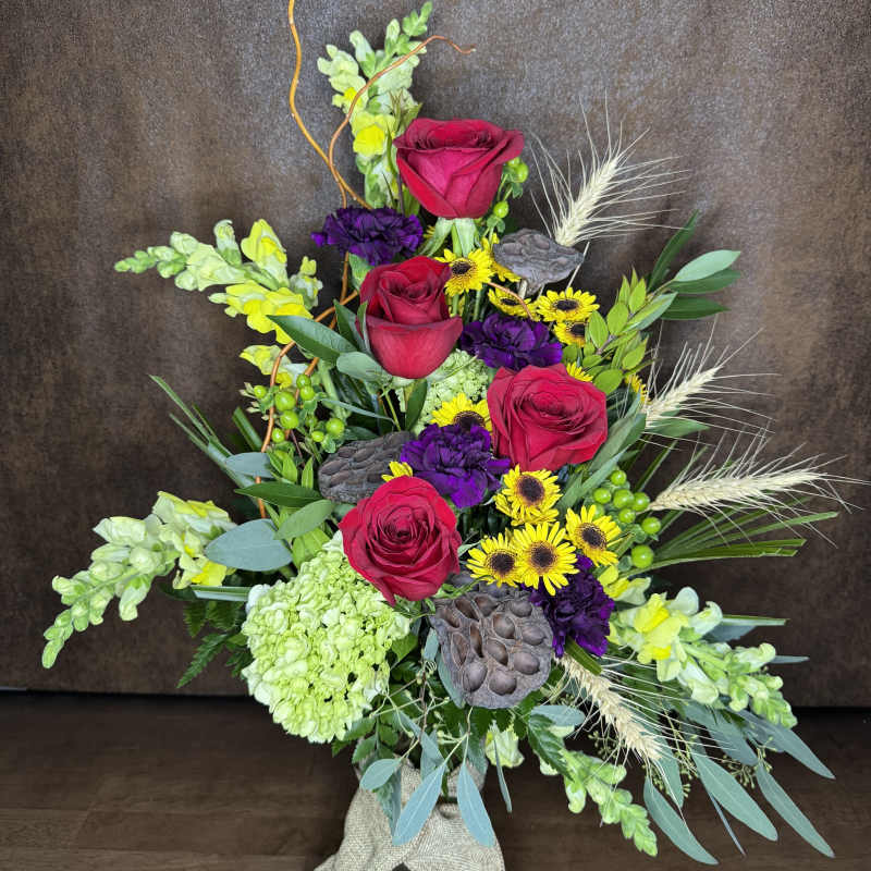 Tall mixed bouquet with red roses, yellow daisies, and purple blooms in a glass vase