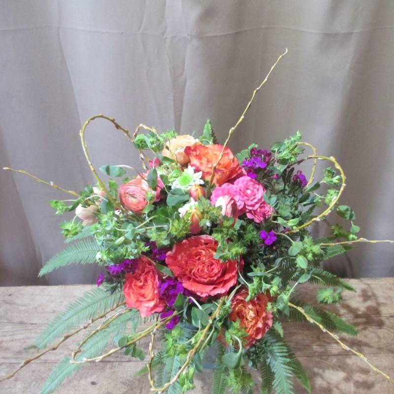 Colorful arrangement of orange and pink roses with purple flowers and curly branches in a low vase