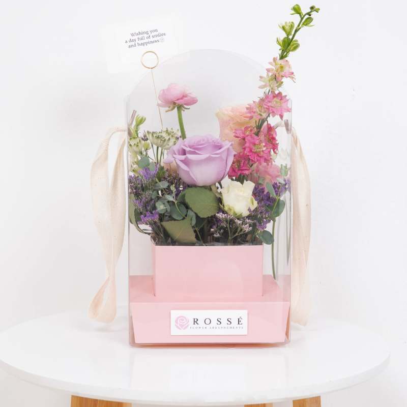 Pastel pink and purple floral arrangement in a pink box with clear cover and small greeting card.