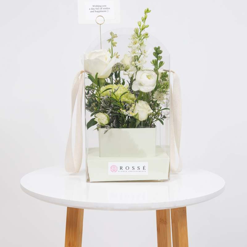 White floral arrangement in a clear gift box with a small card topper
