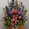 Tall mixed arrangement with pink lilies, pink and yellow roses, and blue delphinium in a white container
