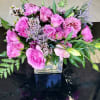 Low arrangement of pink roses and mixed purple flowers in a clear glass cube vase.